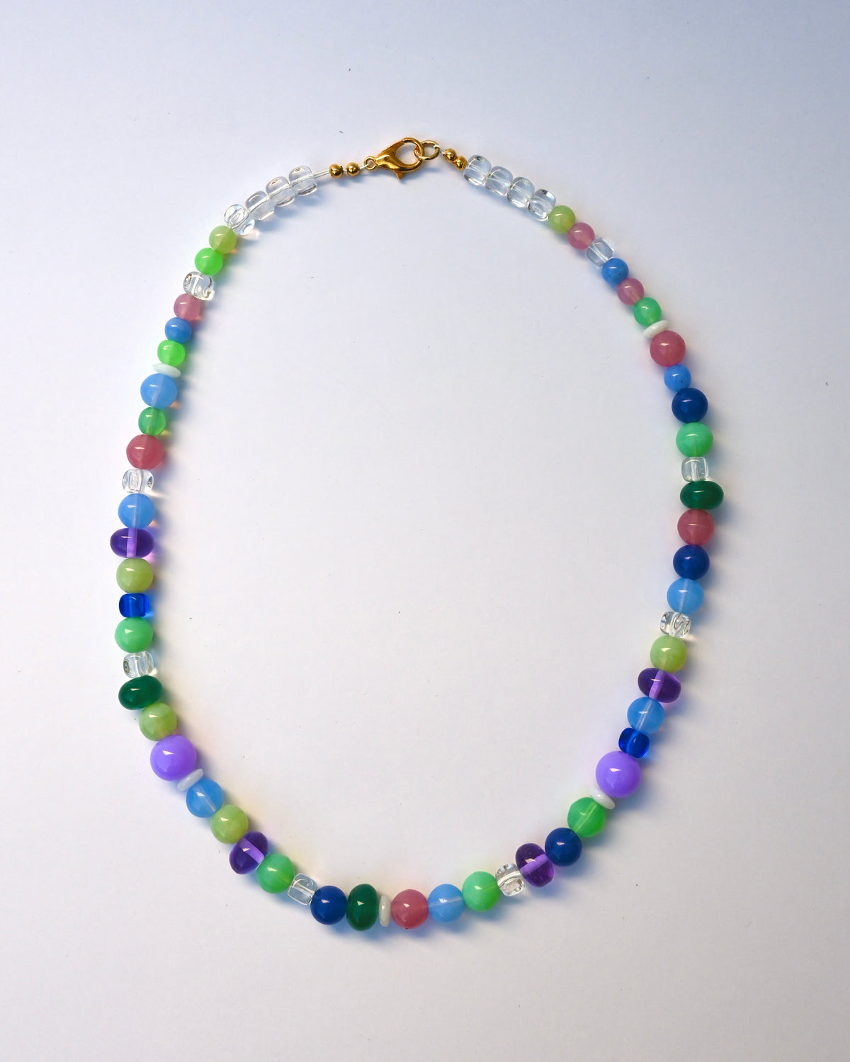 A beaded necklace with multicolored round beads in blue, green, purple, pink, and clear faceted beads, finished with a gold clasp, laid flat on a light gray background.