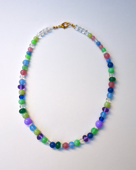 A beaded necklace with multicolored round beads in blue, green, purple, pink, and clear faceted beads, finished with a gold clasp, laid flat on a light gray background.