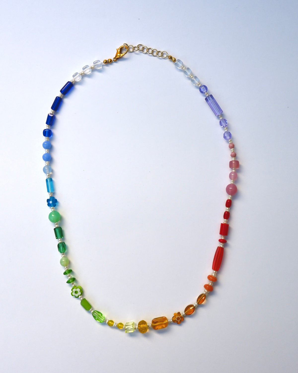 A beaded necklace arranged in an oval shape on a light blue background. The necklace features a rainbow gradient of faceted beads, transitioning from blue and green on the left to yellow, orange, and red on the right, with a gold clasp and chain extender.