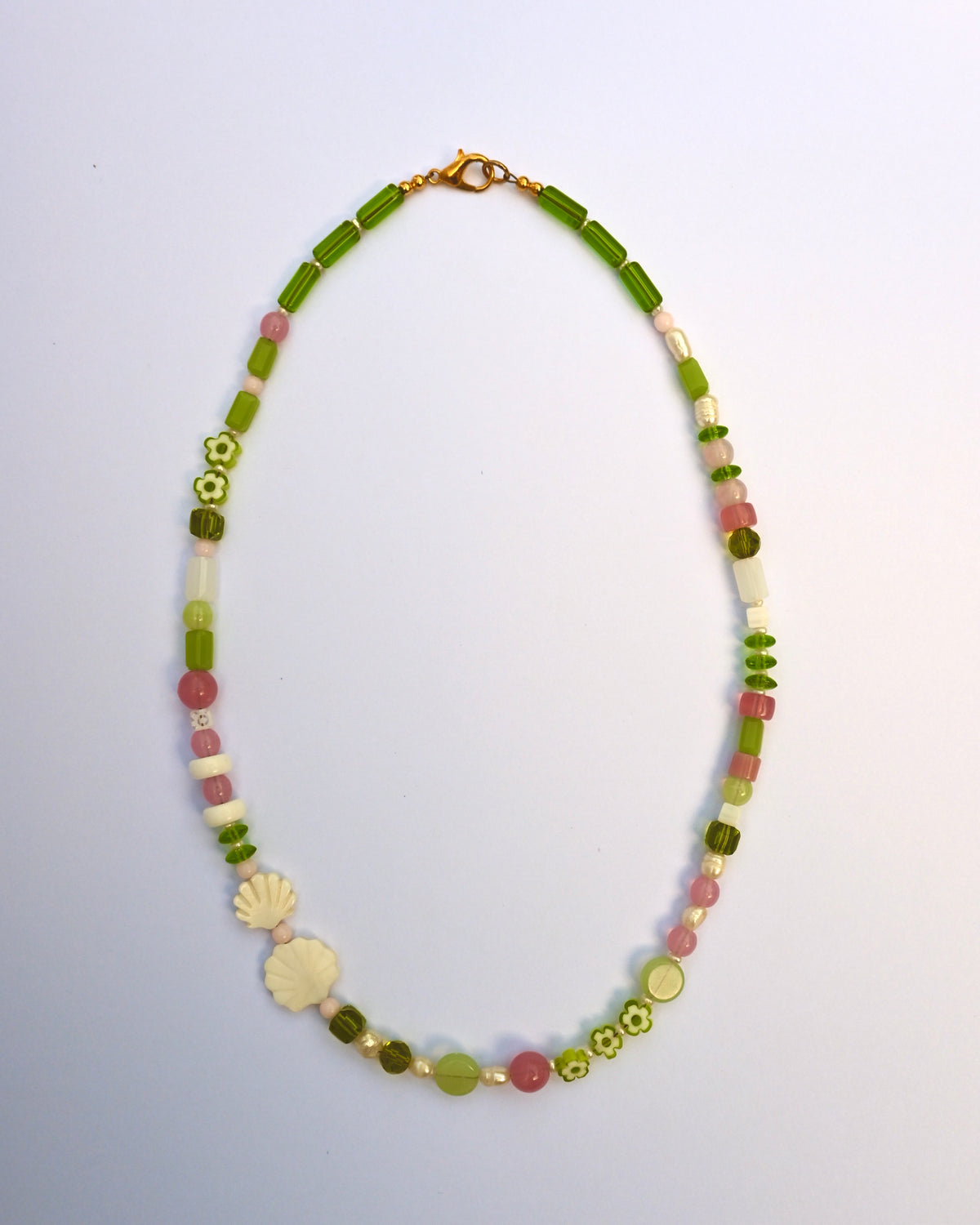 A beaded necklace with green, pink, and white beads, featuring a white shell charm, laid out on a light blue background.