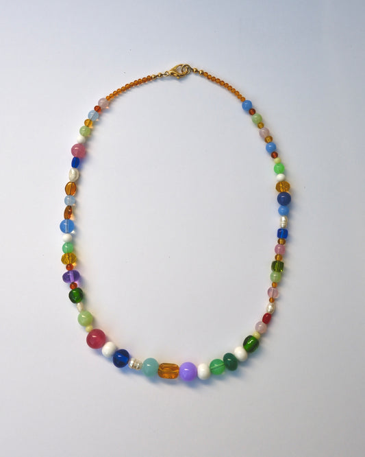 A colorful beaded necklace with a mix of translucent and opaque beads in various colors, featuring a gold clasp, displayed on a light blue background.