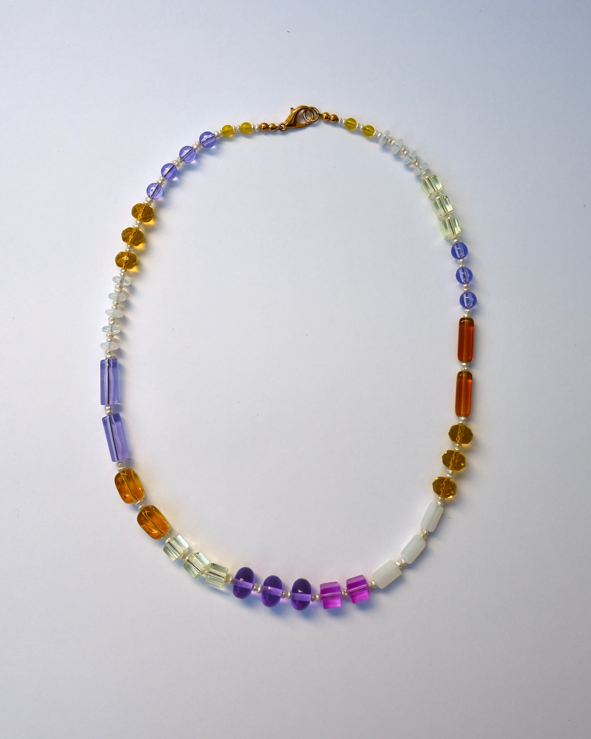 A beaded necklace with a mix of translucent and opaque gemstone-like beads in purple, orange, yellow, white, and clear, arranged in a circle on a light gray background.