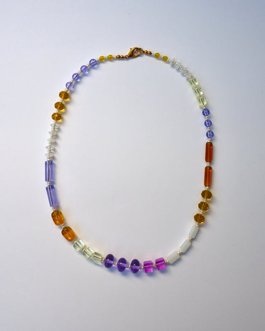 A beaded necklace with a mix of translucent and opaque gemstone-like beads in purple, orange, yellow, white, and clear, arranged in a circle on a light gray background.