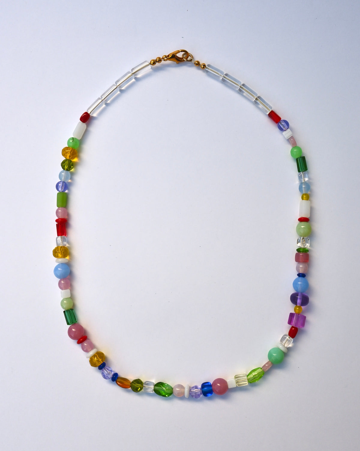A multicolor beaded necklace with a gold clasp, featuring a mix of colorful round and faceted beads in red, green, blue, pink, yellow, and purple, arranged in an oval shape on a light blue background.