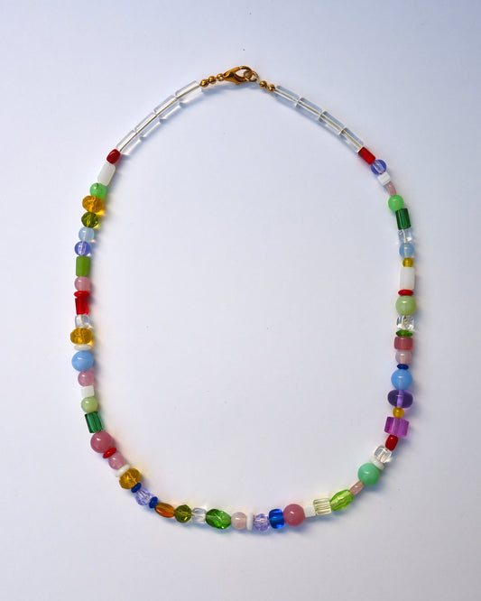 A multicolor beaded necklace with a gold clasp, featuring a mix of colorful round and faceted beads in red, green, blue, pink, yellow, and purple, arranged in an oval shape on a light blue background.